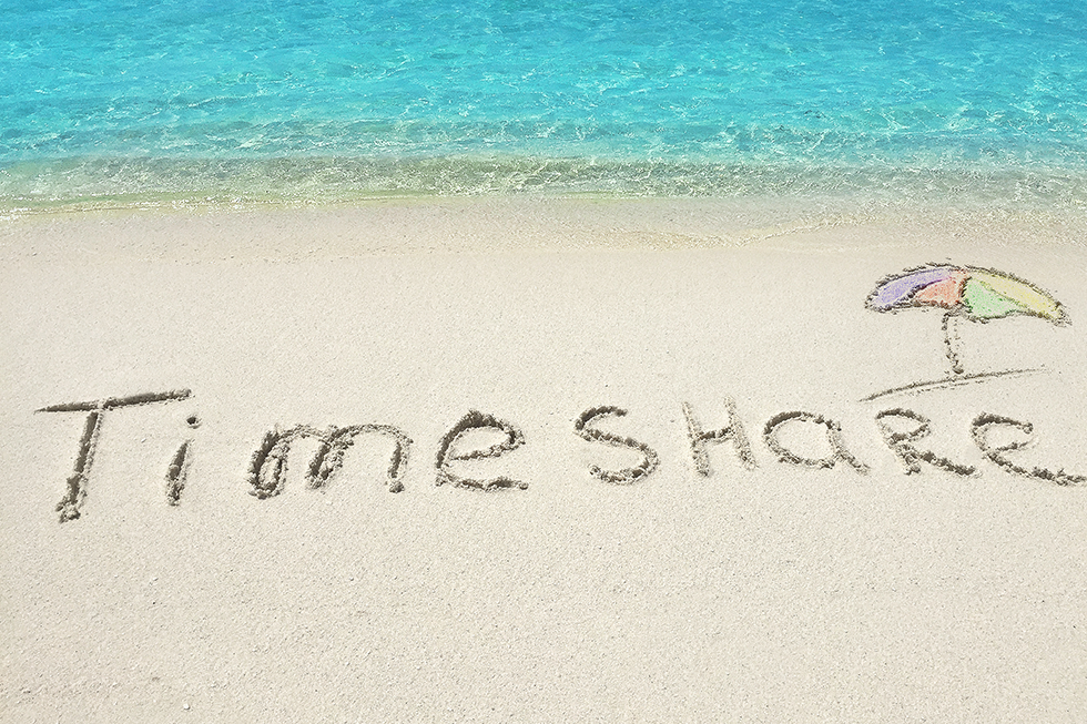 The words "timeshare" are written in the sand at the beach. In the foreground in the sand and in the background is the clear blue water. There is a multi-colored beach umbrella drawn in the sand too.