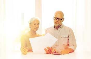 Two old people looking at papers holded by the old woman towards left and the old man towards the right looking at papers holded by woman with hands on calculator placed on the desk