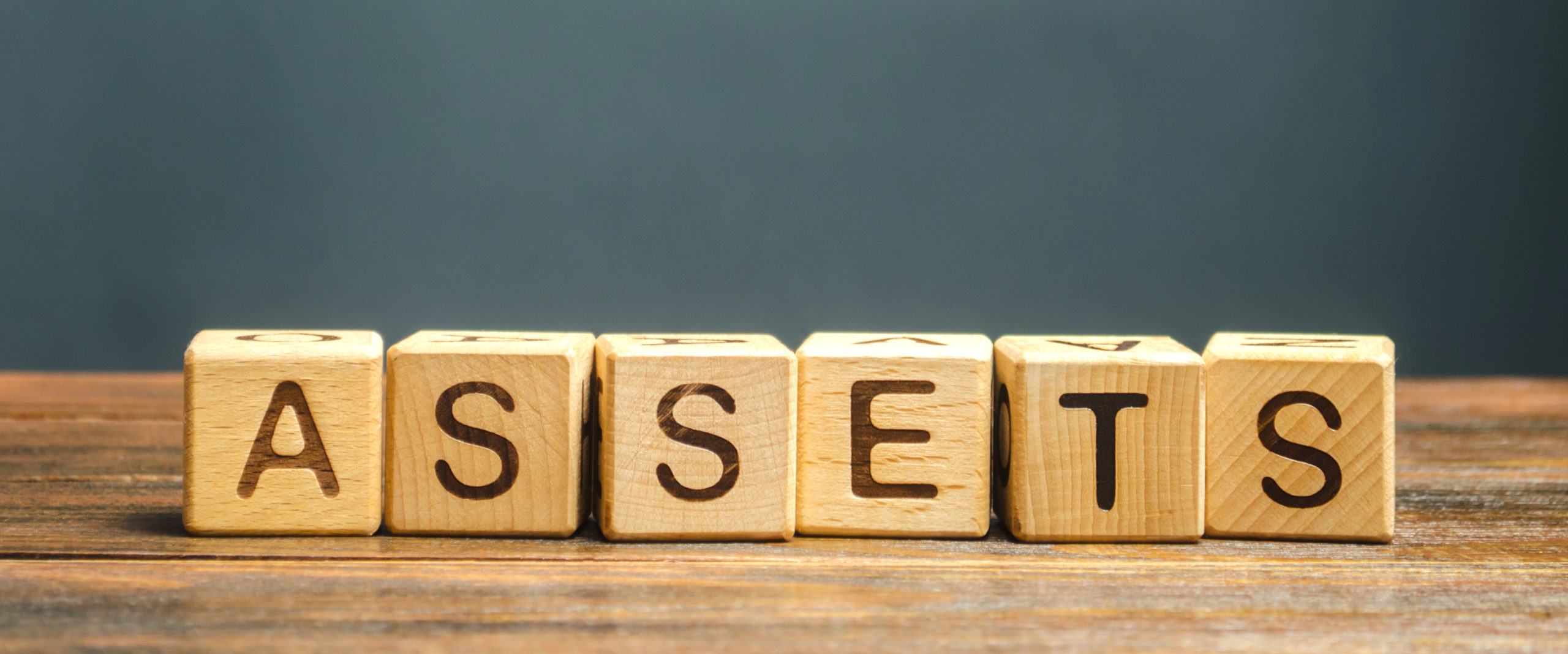 The word "assets" is spelled out in small wooden block letters. The letters sit on a wood surface in front of a dark background.