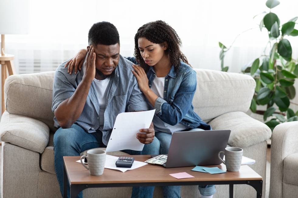 A man and woman sit on a couch. They are The man has a hand on his head as he looks at a paper in front of him. His facial expression suggests that he is distressed. The woman has her arms around him.