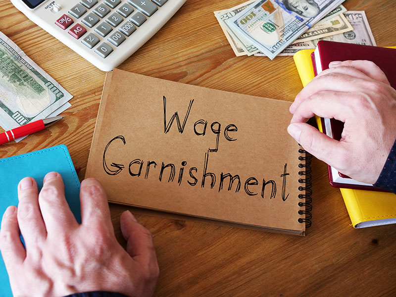A closed brown notebook sits on the table with the words "Wage Garnishment" written on the cover. Two hands are holding either side of the notebook. Around it there are stacks of money, a calculator and other notebooks.