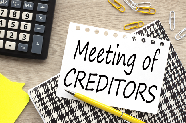 A torn out piece of paper with the words, "Meeting of Creditors," sits on a work desk. Around it are paper clips, a calculator, a pencil and sticky notes.