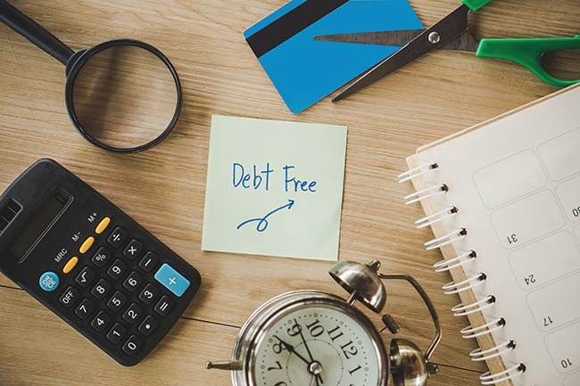 A yellow post-it with the words "Debt Free" in blue ink sits in the center of a wood background. Surrounding the post-it are from the top moving right a credit card, green scissors, a calendar, alarm clock, calculator and a magnifying glass.