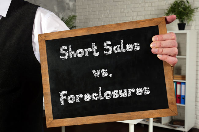 Image shows a person holding a chalk board with one hand. On the chalkboard it says, "Short Sales vs. Foreclosures." We can see half of the person's torso. They are wearing a long sleeve white shirt and black buttoned vest.