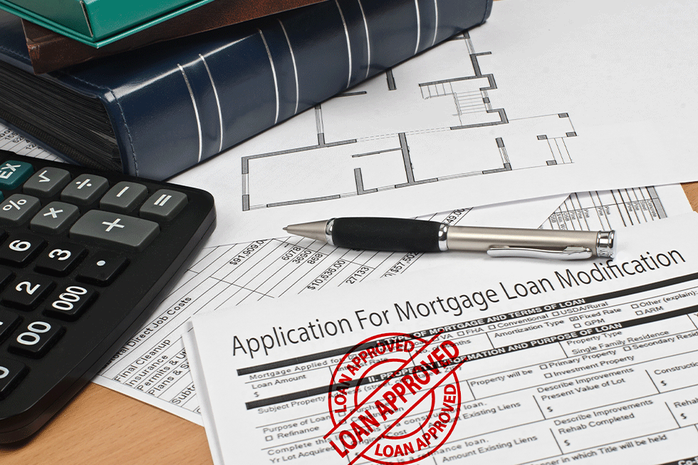 Papers on a desk with a pen, calculator and a small stack of books. The top paper says, "Application For Mortgage Loan Modification" and it has a red stamp on it that says, "Loan Approved."