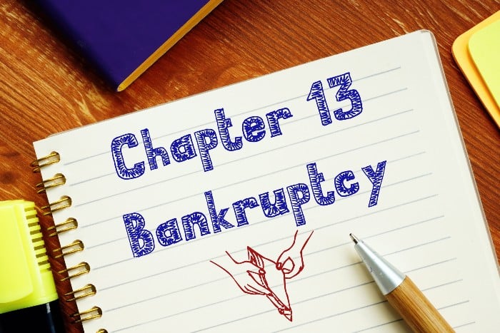 A notebook sits open on a wooden table. The words, "Chapter 13 Bankruptcy" are written in pen. Underneath is a doodle of hands opening up a wallet.