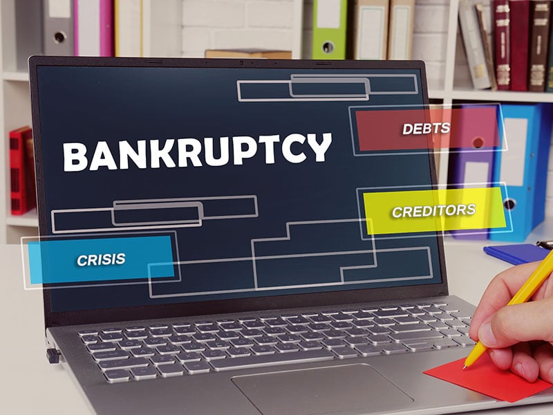 The words "Bankruptcy," "Debts," "Creditors" and "Crisis appear in front of a laptop screen. A hand in the bottom right corner is writing on a red sticky note with a yellow pen. No writing is visible.