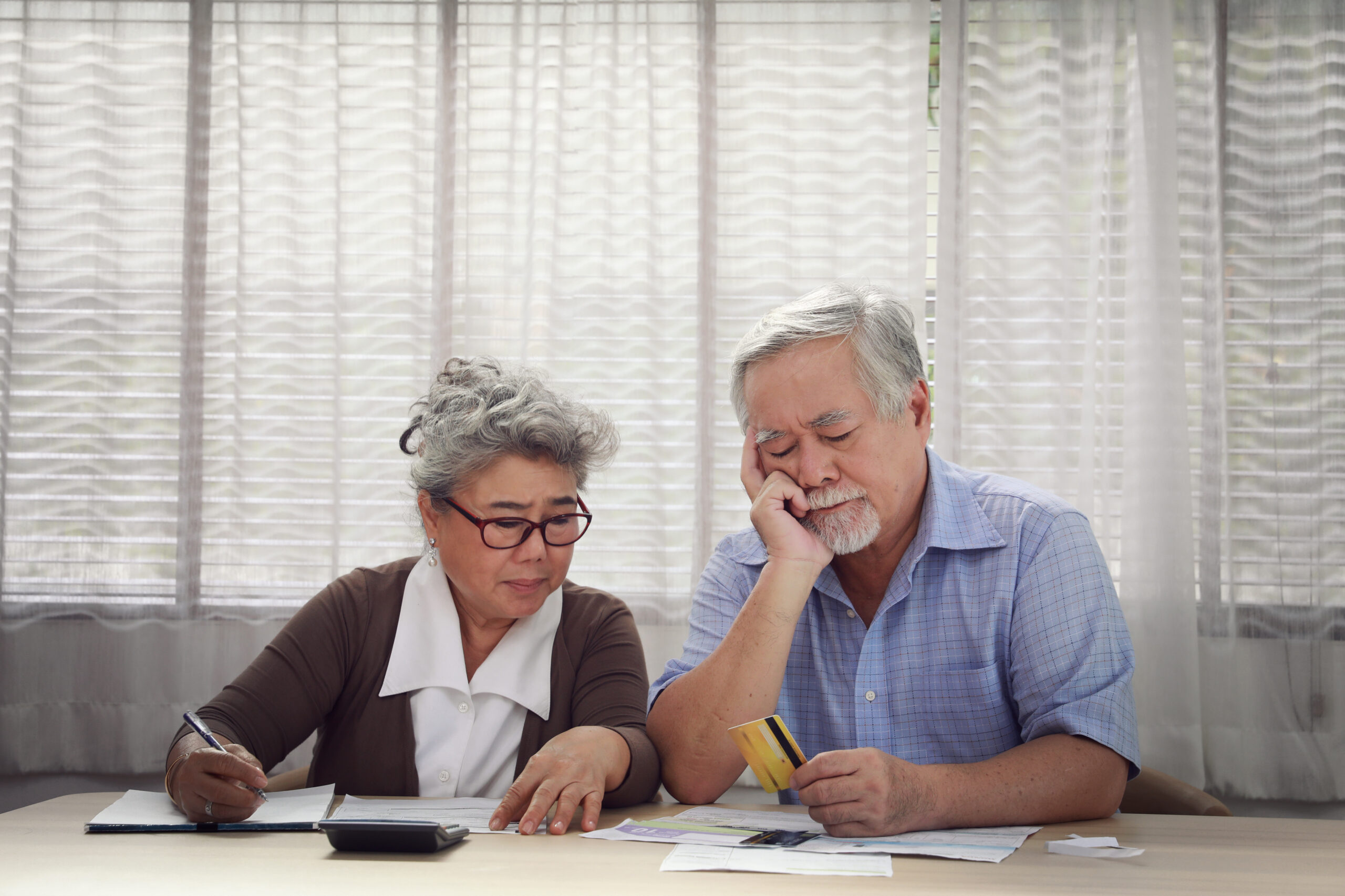 A man and a woman sit at a table in front of curtain covered windows. They look down despondently at a credit card and table full of bills and receipts.